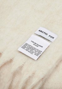Écharpe beige clair avec une texture douce et duveteuse. Les étiquettes indiquent "ARCTIC FOX" et "L'ÉCHARPE DE REYKJAVIK," avec des instructions d'entretien en texte noir.
