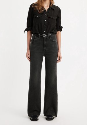 Woman standing wearing black button-up shirt with rolled sleeves, high-waisted black wide-leg jeans, black belt, and black shoes.