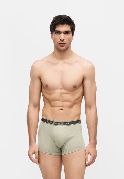 Μυώδης άνδρας όρθιος, φορώντας μπεζ boxer Emporio Armani, με κοντά σκούρα μαλλιά και ουδέτερη έκφραση προσώπου σε λευκό φόντο.