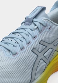 Κοντινή λήψη ανοιχτό μπλε παπουτσιού τρεξίματος Asics με υφή υφάσματος, κορδόνια με μοτίβο, γκρι λογότυπο και κίτρινη σόλα με διαβάθμιση χρώματος.