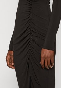 Czarna sukienka z elastycznego materiału, z długimi rękawami i skomplikowanym pionowym marszczeniem po bokach, podkreślającym teksturowane detale.