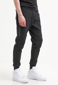 Man som bär svarta tapered joggerbyxor med dragsko och vita högsnörade sneakers, stående framför en helt vit bakgrund.