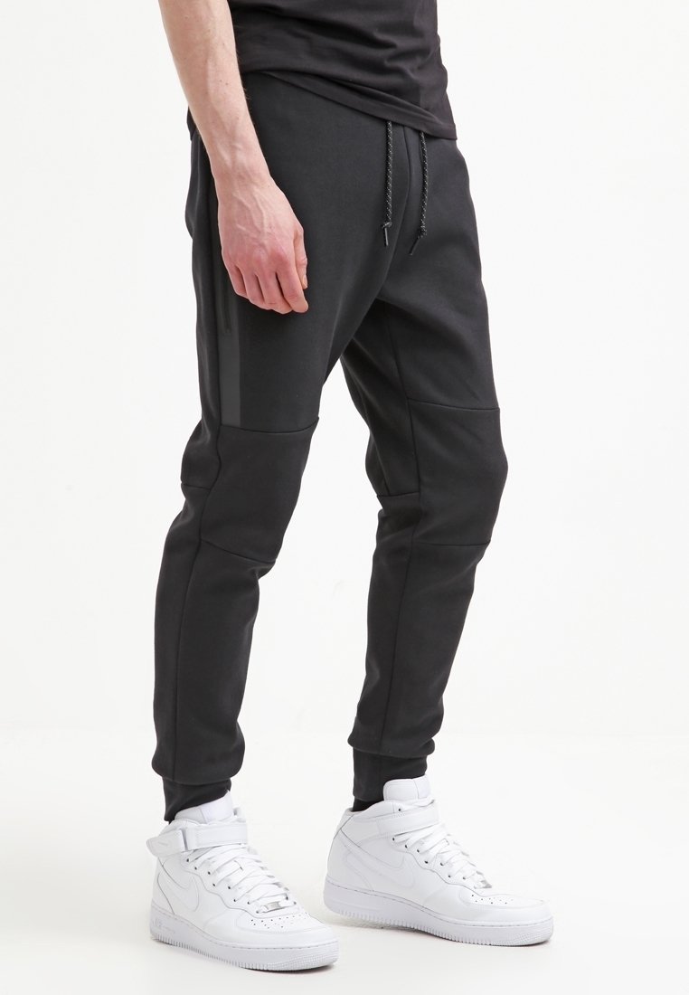 Man som bär svarta tapered joggerbyxor med dragsko och vita högsnörade sneakers, stående framför en helt vit bakgrund.