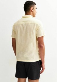 Camicia gialla chiara a maniche corte con colletto e una texture liscia. Indossata con pantaloni corti neri a motivo, vista da dietro.