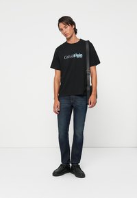 Čierne bavlnené tričko s kontrastným modrým logom "Calvin Klein", v kombinácii s modrými džínsami a čiernymi teniskami. Model stojí s ramennou taškou.