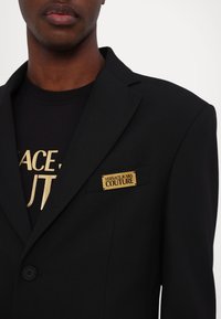 Blazer nero su misura con rever a punta, featuring un distintivo in smalto dorato "Versace Jeans Couture" sulla tasca. Tessuto testurizzato.