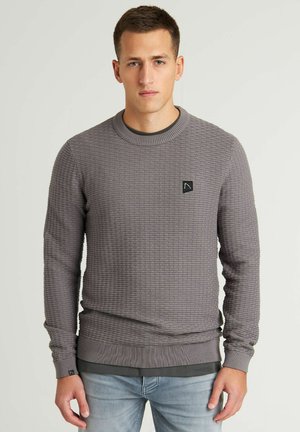 Grau strukturierter Pullover mit geripptem Kragen und Bündchen, ausgestattet mit einem kleinen schwarzen Logo-Patch. Getragen über einem dunklen Hemd.