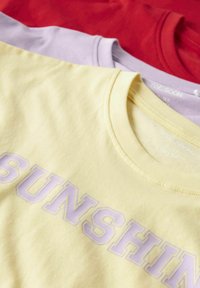 Tre t-shirts staplade: gul med "SUNSHINE" i lavendel, ljuslila och röd. Baumwollmaterial, rund hals, slät textur.