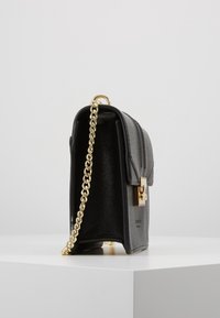 Borsa a mano in pelle nera con finitura testurizzata, dotata di tracolla in catena dorata e chiusura quadrata in oro, posta in posizione verticale su una superficie bianca e con uno sfondo grigio.