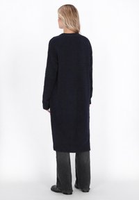 Langer marineblauer Strickpullover mit lockerer Passform, gerippten Bündchen und mittlerer Oberschenkellänge, kombiniert mit dunkelgrauen, weiten Hosen und schwarzen Schuhen.