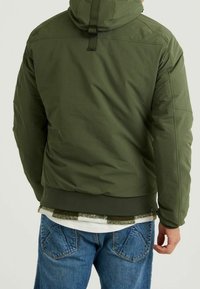 Olivově zelená zateplená bunda s kapucí, se stahovací šňůrou, žebrovaným lemem a prošívanou strukturou. Nosená přes bílo-zelenou kostkovanou halenku.