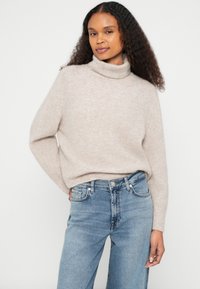 Camisola de gola alta bege clara em material de malha suave, corte largo com ombros caídos, combinada com jeans de ganga azul claro.