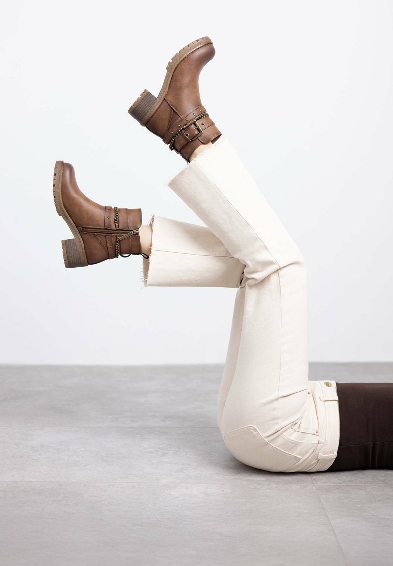 Botas de cuero marrón hasta el tobillo con un tacón apilado, que presentan dos correas y hebillas decorativas, combinadas con jeans de color claro y deshilachados.