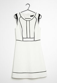 Bezrukávové bílé šaty s černými geometrickými liniemi, kulatým výstřihem, strukturovanou tkaninou a zipovým zapínáním na zádech. Visí na černém háčku.