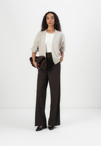 Light beige cardigan, white top, high-waisted brown trousers, black pointed heels. Model holds a dark brown clutch, featuring soft textures.