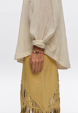 Brazo con una blusa crema remangada y una falda amarilla con flecos, adornado con una pulsera de cuentas de colores en la muñeca.