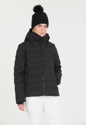 Kvinde iført en sort dunjakke med hætte og en sort strikket hat med pompon, kombineret med hvide bukser mod en ensfarvet baggrund.