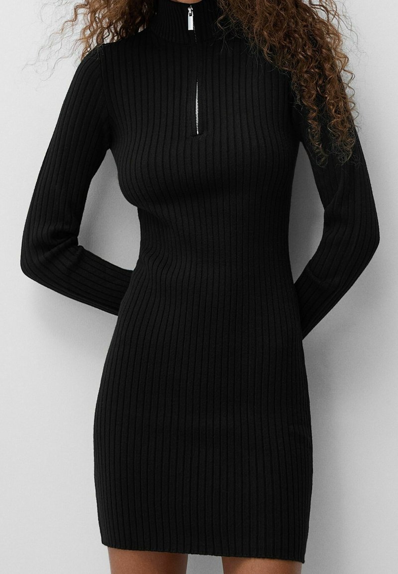 Černé žebrované pletené šaty s vysokým límcem a polovičním zipem, obepínající střih a dlouhé rukávy.