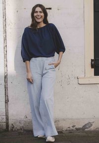 Navy-blauwe top met ruime mouwen en een bootnek, gecombineerd met lichtblauwe gestreepte wijde broek. Model staat tegen een gestructureerde muur.