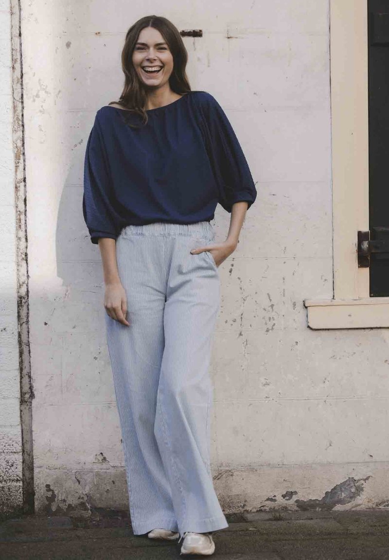 Navy-blauwe top met ruime mouwen en een bootnek, gecombineerd met lichtblauwe gestreepte wijde broek. Model staat tegen een gestructureerde muur.