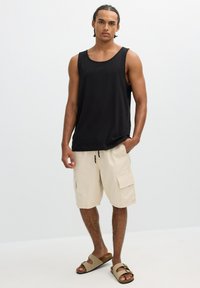 Joven de pie con camiseta negra sin mangas, pantalones cortos cargo beige y sandalias beige sobre un fondo claro y uniforme.