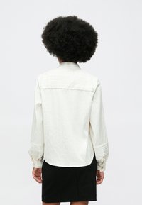 Mulher com cabelo afro usa uma blusa branca de manga longa e uma saia preta, vista de costas contra um fundo simples.