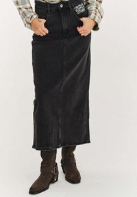 Čierna rifľová maxi sukňa s priamym strihom, prednými vreckami a dekoratívnym prešitím v oblasti pása. Kombinovaná s hnedými semišovými čižmami.