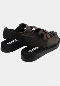 Brown sandals with a thick black sole, featuring a textured strap and elastic band. 'CAMPER' logo in white on the footbed.