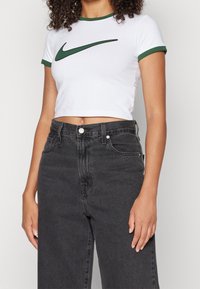 Valkoinen lyhyt t-paita, jossa on vihreä reunus ja suuri musta Nike Swoosh -logo, yhdistetty korkeavyötäröisiin mustiin farkkuihin.