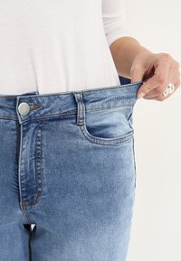Blå denimjeans med en avslappnad passform, som har knappstängning, fem fickor och lätt slitage. Hålls upp av en hand som visar överflödig midjeband.