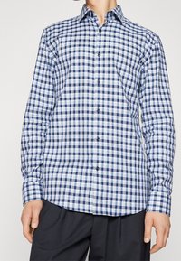 Uomo che indossa una camicia a maniche lunghe aderente con un motivo a scacchi blu e bianco e pantaloni neri, in piedi di fronte a uno sfondo semplice.