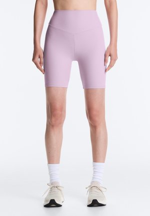 Nő, aki magas derekú levendulalila biciklis rövidnadrágot, fehér zoknit és bézs sportcipőt visel, karjai lazán a teste mellett állnak fehér háttér előtt.
