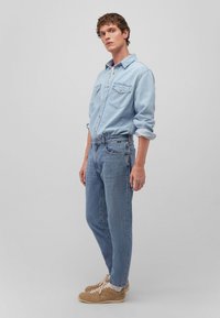 Camicia di jeans blu chiaro con due tasche sul petto, abbinata a jeans blu chiaro e sneakers beige, in una posa in piedi su uno sfondo neutro.