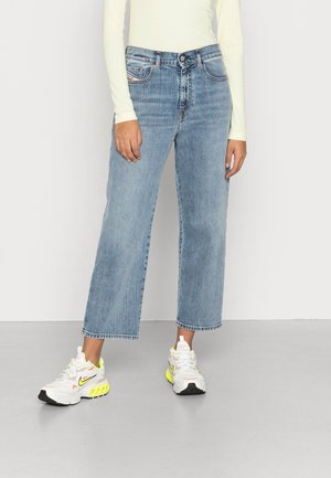 Persoon die lichtblauwe cropped straight-leg jeans draagt, een wit top met lange mouwen en witte sneakers met gele en zwarte details, staand op een grijze vloer.