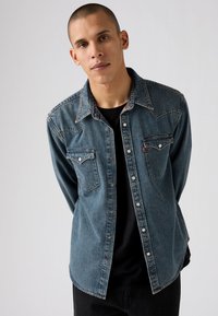 Cămașă din denim albastru cu guler ascuțit, două buzunare pe piept și capse. Prezintă o textură estompată și detalii de cusut în contrast.