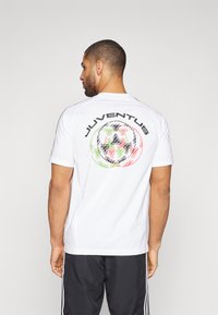Biała koszulka z krótkim rękawem z okrągłym, wielokolorowym nadrukiem oraz wyraźnym napisem "JUVENTUS" umieszczonym na plecach.