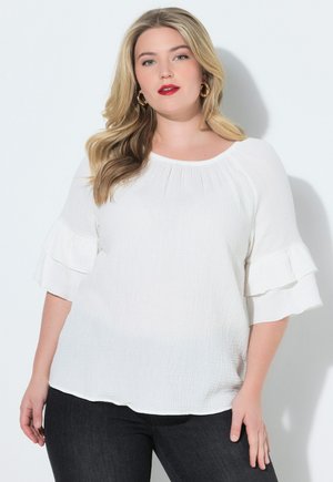 Frau mit langen blonden Haaren, die goldene Creolen, eine weiße Bluse mit Struktur und Rüschenärmeln sowie dunkle Jeans trägt, vor hellem Hintergrund.