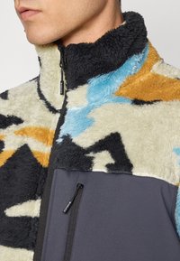 Giacca in pile con motivo astratto multicolore in beige, blu, arancione e nero. Presenta un colletto alto e una tasca con zip frontale.