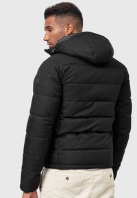 Schwarze Pufferjacke aus strukturiertem Stoff, mit Kapuze und gestepptem Design. Sie hat einen Schulteraufnäher und wird über einer hellen Hose getragen.