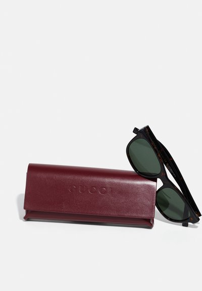 Ochelari de soare tortoiseshell negri cu lentile verzi, alături de un etui din piele burgundă închisă, embosat cu "GUCCI".