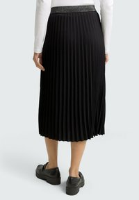 Jupe midi plissée noire avec une taille texturée, tombant en dessous du genou. Associée à des chaussures noires, mettant en avant le design fluide de la jupe.