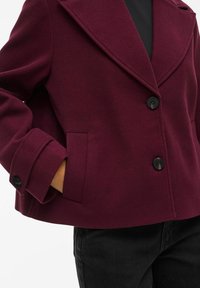 Burgundowa kurtka z mieszanki wełny z wykładanym kołnierzem, czarnymi guzikami i bocznymi kieszeniami. Posiada zdefiniowaną sylwetkę i miękką fakturę.