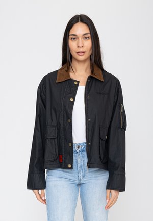 Frau mit langen dunklen Haaren trägt eine schwarze Jacke mit braunem Kragen über einem weißen Hemd und hellblauen Jeans, steht vor einem schlichten Hintergrund.