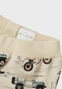 Beige Baumwollhosen mit einem gerippten Bund und einem Knopfverschluss, verziert mit Fahrzeugmustern in Braun- und Schwarztönen.