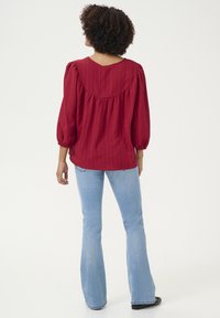 Red ribbed blouse with a round back yoke, puff sleeves, and a loose fit. Paired with light blue flared jeans and black shoes.