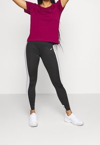 Camiseta burdeos con hombros caídos, combinada con unas mallas negras que tienen franjas laterales blancas y un pequeño logo, completado con zapatillas blancas.