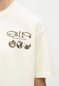 Beige T-skjorte med teksten «Nature in Motion», abstrakte former og tre illustrasjoner av jordkloden på øvre del av brystet.