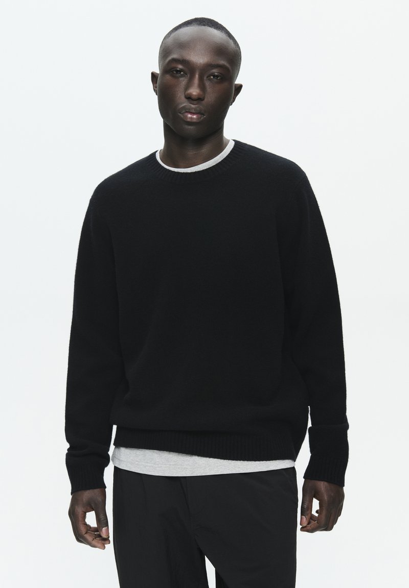 Juodas megztinis su apvalia iškirpte ir raštuotais rankogaliais, uždėtas ant pilkos marškinėlių. Lygi tekstūra, laisvas siluetas ir minimalus dizainas.