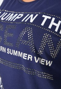 Navig blauwe t-shirt met witte tekst, versierd met zilveren rhinestones in verschillende patronen. Korte mouwen, zachte textuur en een casual pasvorm.