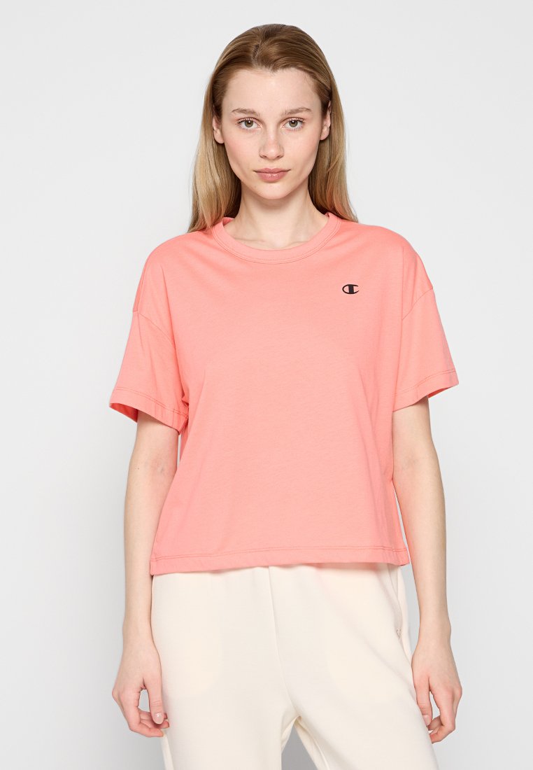 Jeune femme aux cheveux blonds raides portant un t-shirt oversize corail avec un petit logo sur la poitrine et un pantalon large crème.